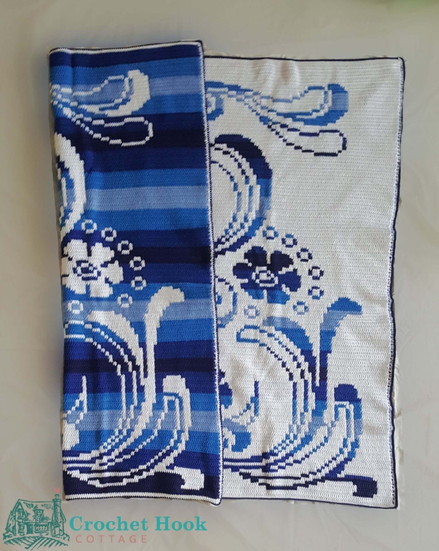 Crocheted Art Nouveau Blanket featuring a sylized pedestal adorned with flowers and flourishes in ombre blue and white, the blanket is partly folded over to show how the design is the same on both sides of the blanket but the colours are reversed