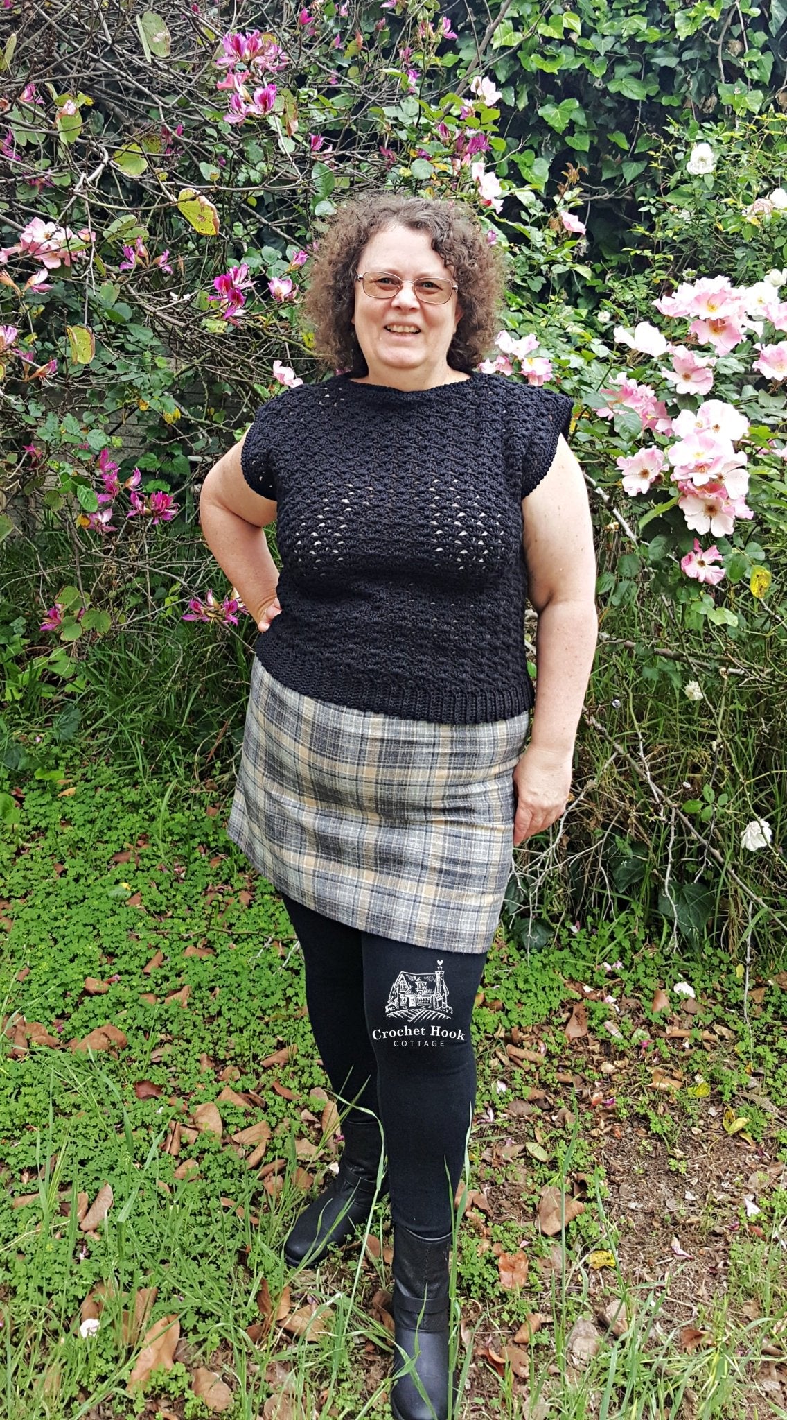 person wearing the black Shell-patterned Sleeveless Ladies Top, size ladies medium, with a tartan skirt, black leggings and boots, standing in front of a rose bush - www.crochethookcottage.com