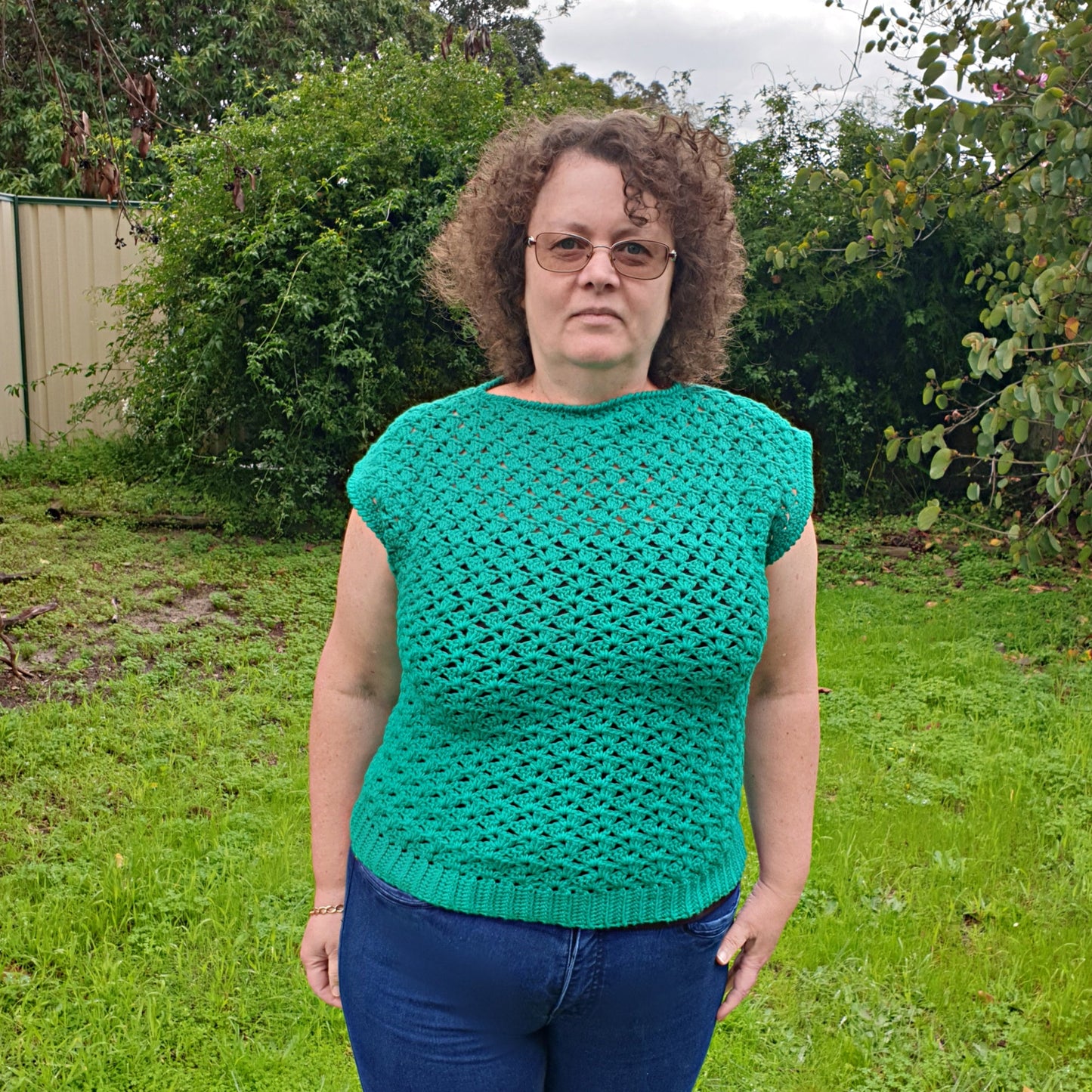 person wearing the green Shell-patterned Sleeveless Ladies Top, size ladies medium, a pair of jeans, outside on a patch of grass with bushes behind - www.crochethookcottage.com